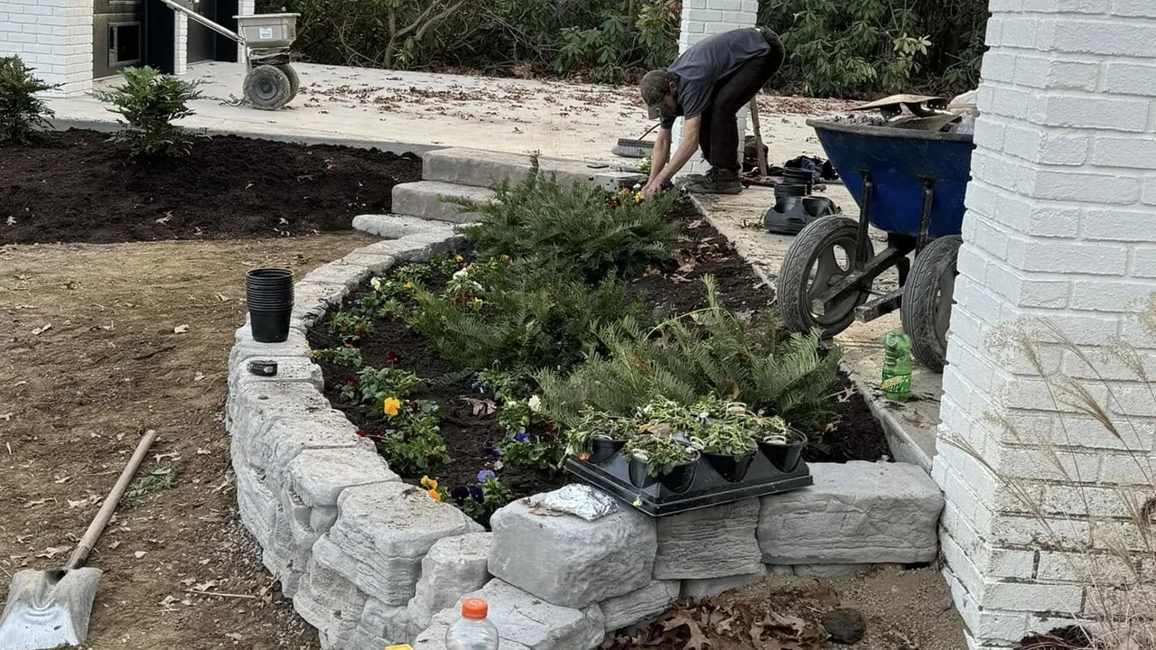 Landscaping crew building a stone block wall and planting bed beside a white home