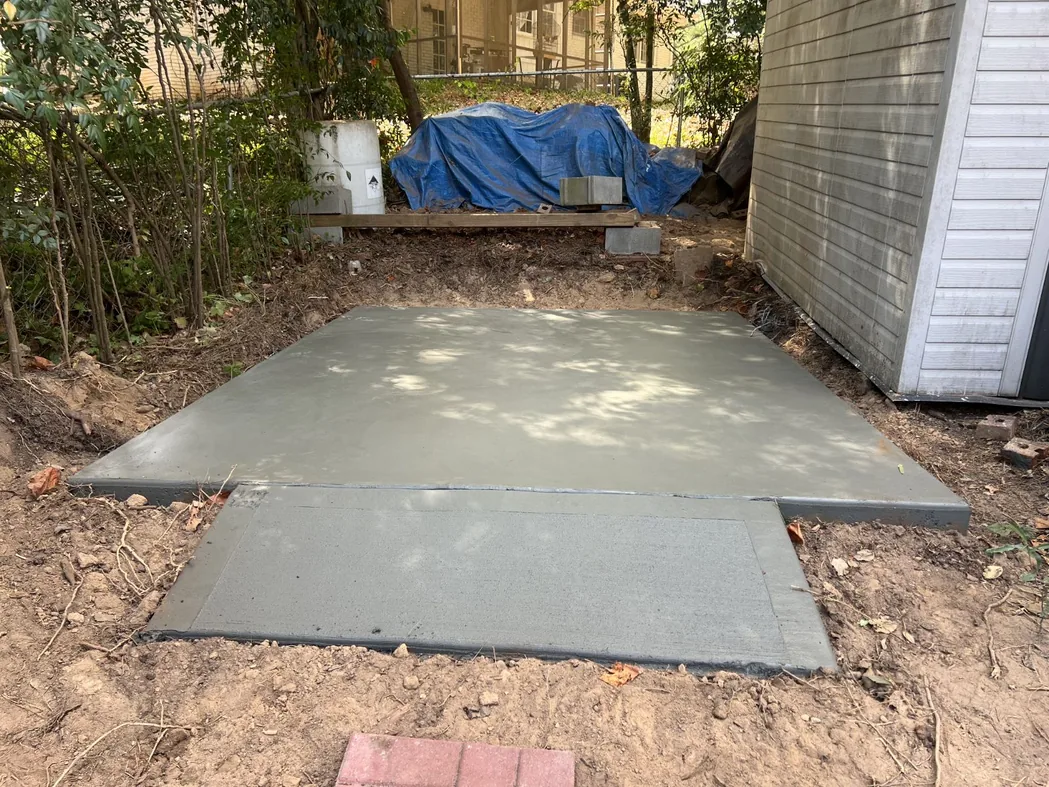 Concrete: a freshly poured plain concrete shed pad with a small apron slab beside a vinyl sided shed