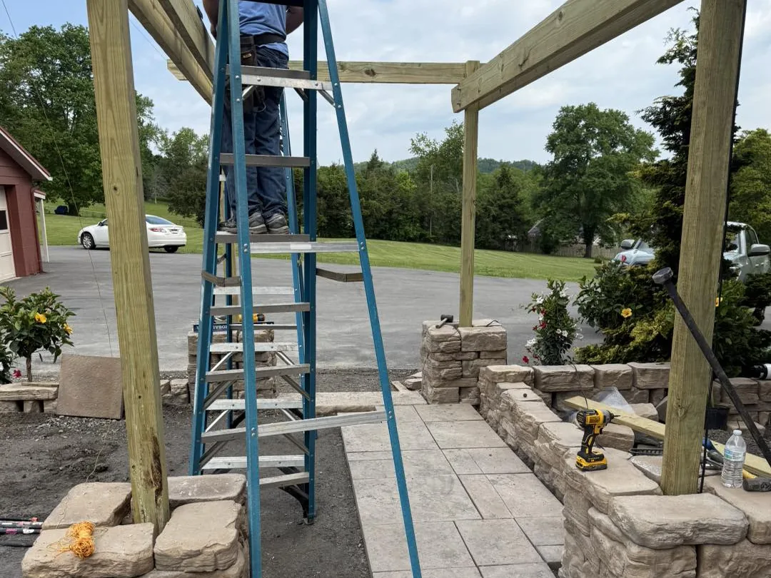 Hardscaping: a crew member on a ladder assembling a wood arbor over a paver walkway with stone pillars