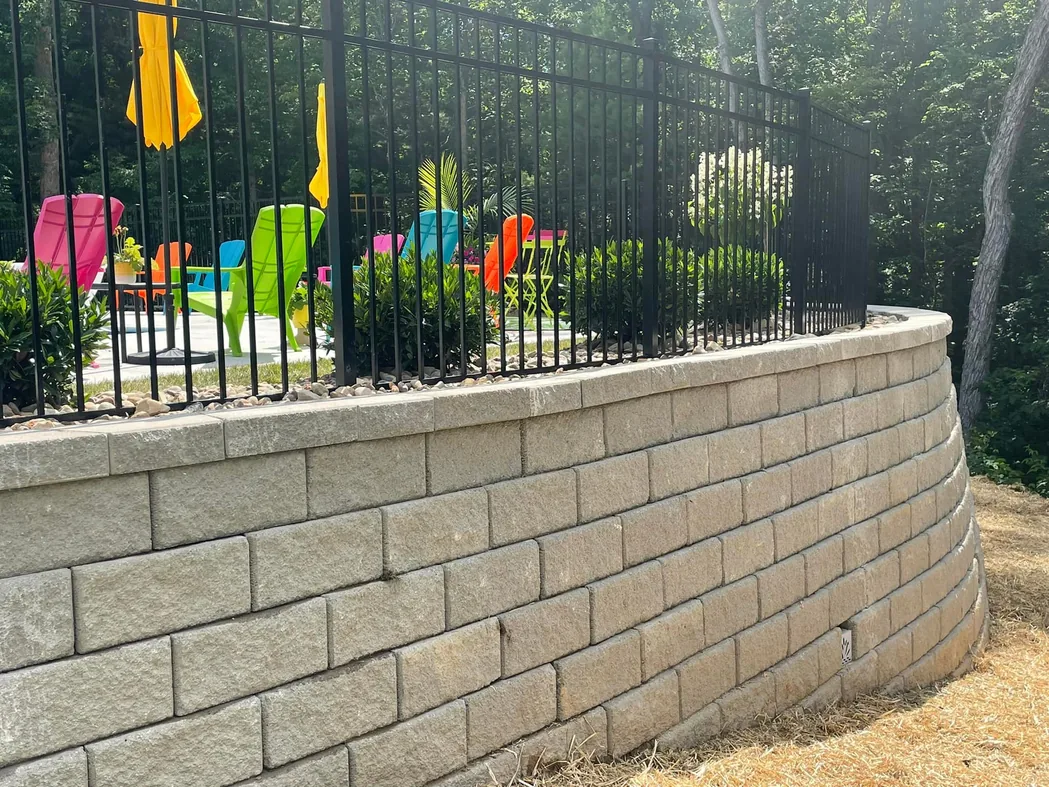 Hardscaping: a tall curved block retaining wall below a fenced patio with bright lawn chairs