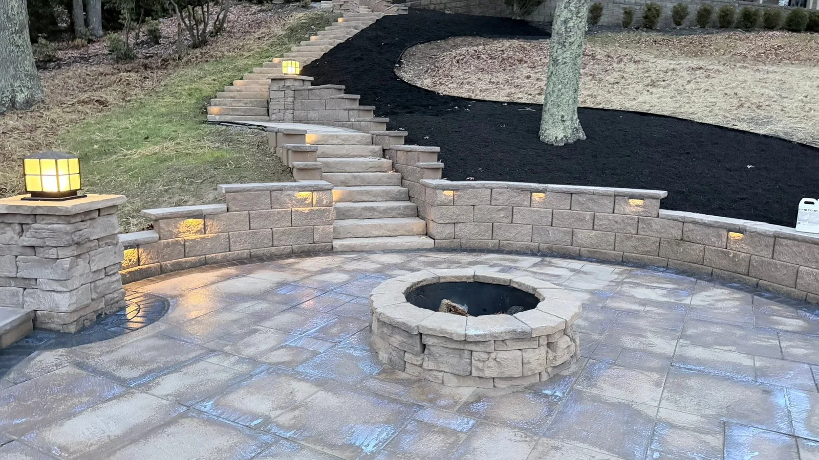 Landscaping and hardscaping: a paver patio with a stone fire pit, lighted seating walls and steps rising through fresh mulch beds