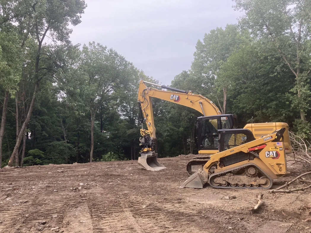 Land clearing: an excavator and a tracked loader on a freshly cleared dirt lot ringed by hardwood trees