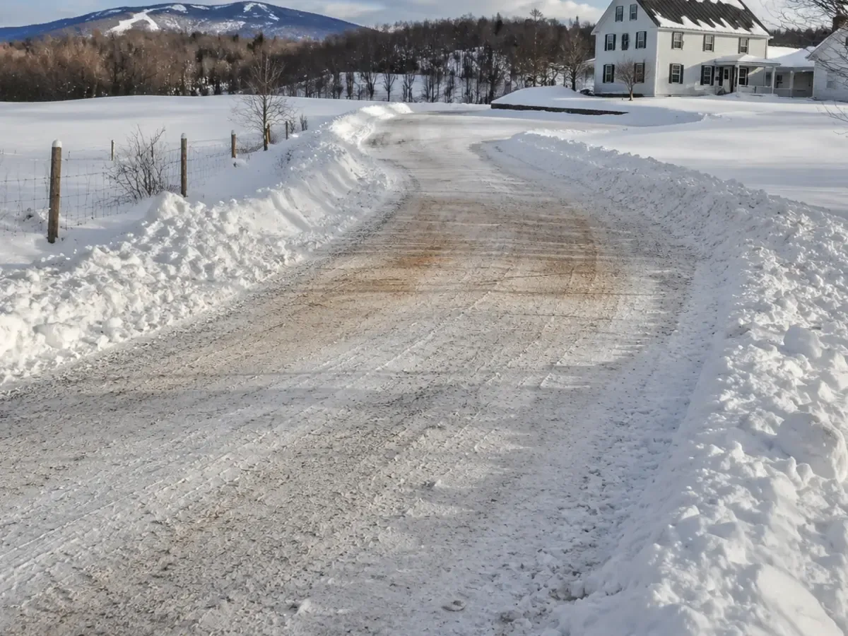 Snow removal: a plowed rural gravel driveway between snowbanks leading to a farmhouse with wooded hills behind