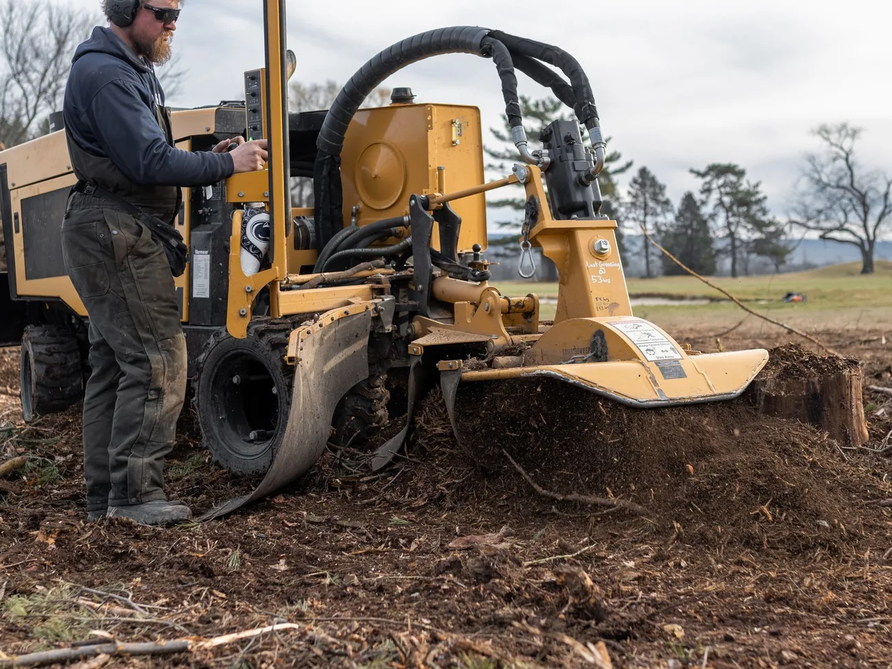 Stump grinding: an operator running a self propelled stump grinder on a cleared lot with chips flying and trees behind