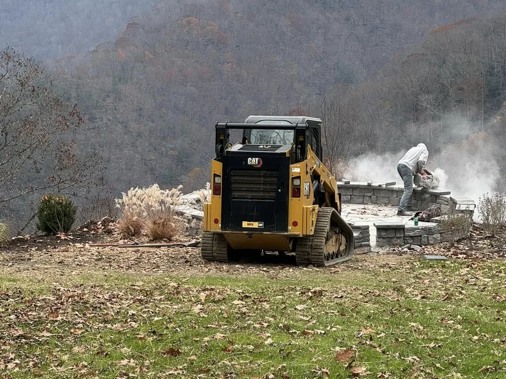 Tree removal: a tracked skid steer on a hillside job with wooded mountains behind